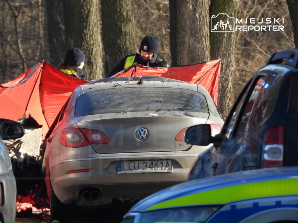 Mężczyzna w mundurze policyjnym trzyma czerwoną płachtę, obok stoi zaparkowany samochód osobowy w lesie.