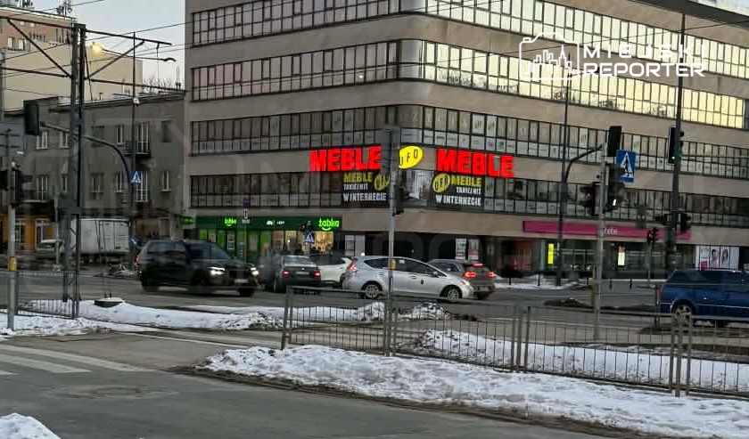 Nowoczesny budynek z przeszkloną fasadą, na parterze widoczny neon "MEBLE", wokół ruch uliczny i śnieg.