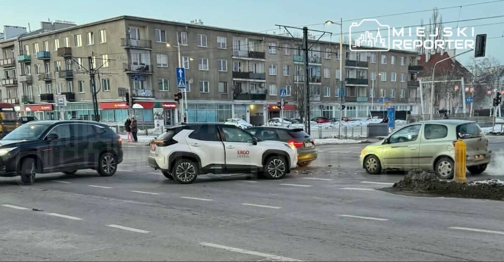 Dwa samochody osobowe zderzone na skrzyżowaniu, w tle nowoczesne budynki mieszkalne i sklepy.