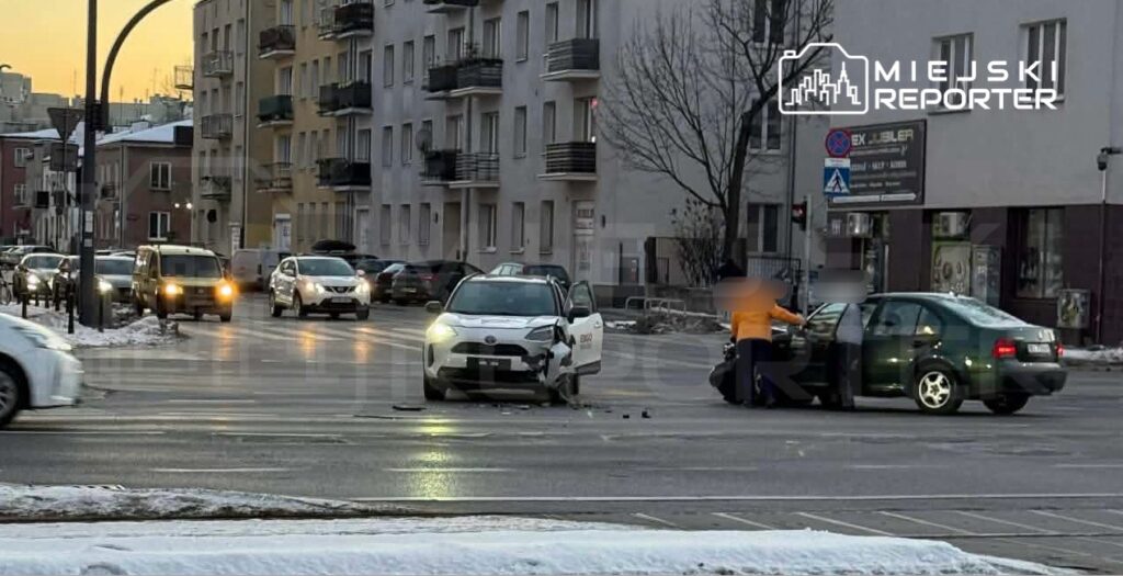 Dwa uszkodzone samochody stoją na skrzyżowaniu, a kierowcy rozmawiają obok pojazdów. W tle widać bloki mieszkalne.