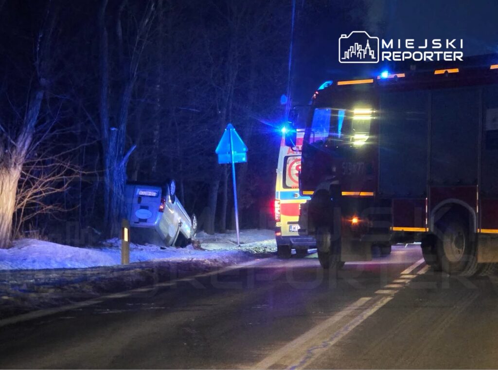 Samochód osobowy leży na dachu przy drodze, obok stoją strażacy i karetka z włączonymi sygnałami świetlnymi.