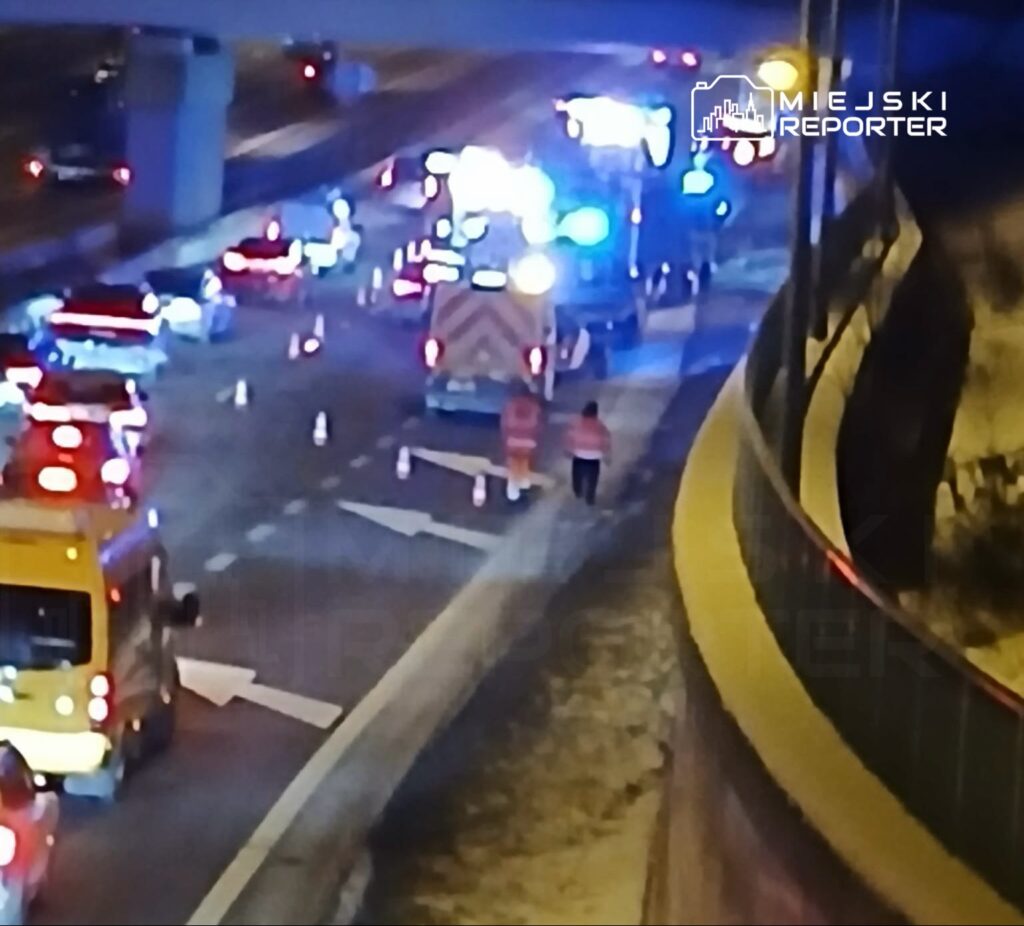 Na drodze widać pojazdy służb ratunkowych oraz kilka osób w odblaskowych kamizelkach, pracujących przy wypadku.