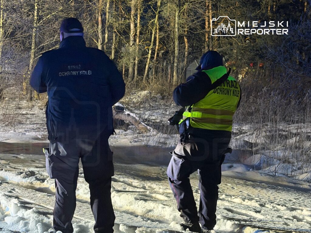 Dwaj mężczyźni w odzieży służbowej Ochrony Kolei stoją na zaśnieżonym terenie, obserwując otoczenie.