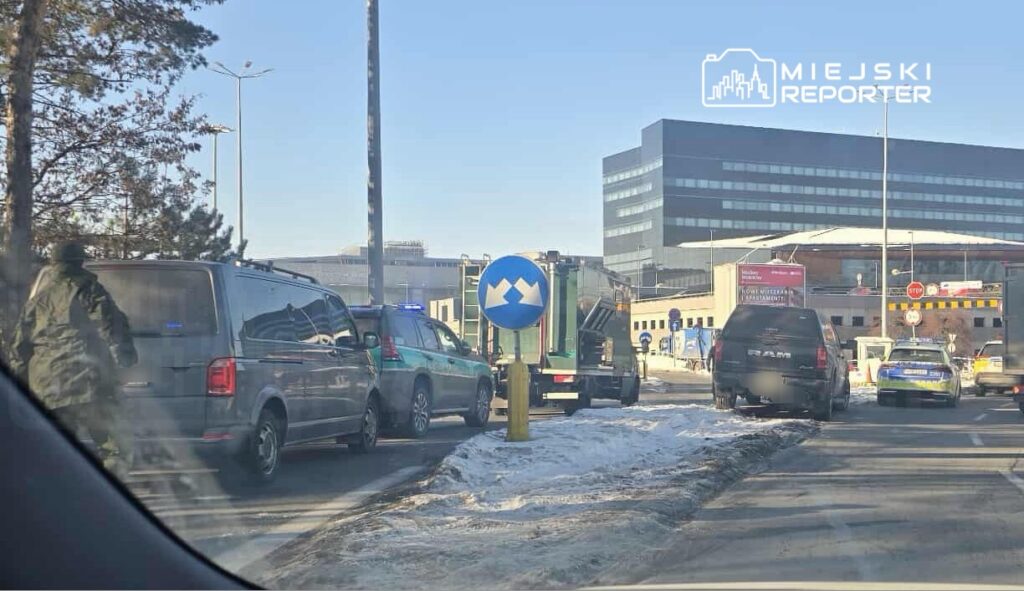 Grupa pojazdów, w tym furgonetki i samochody policyjne, zatrzymana na drodze obok nowoczesnego biurowca.