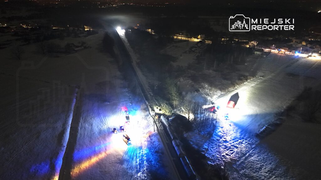 Na zaśnieżonym terenie w nocy, pojazdy ratunkowe oświetlają tory kolejowe, gdzie doszło do wypadku.