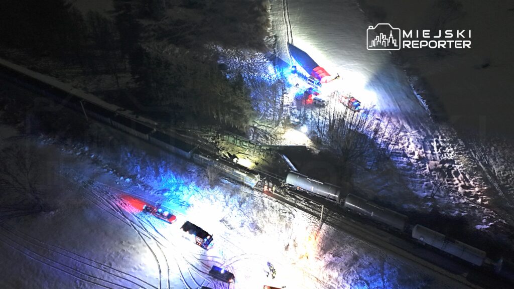 W nocy, na ośnieżonym terenie, ekipy ratunkowe pracują przy wykolejonym pociągu, oświetlając miejsce zdarzenia.