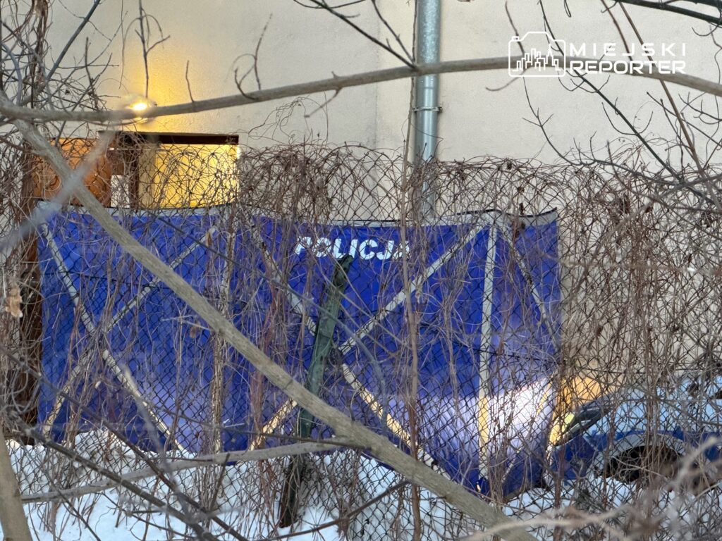 Nieoznakowany samochód policyjny stoi za ogrodzeniem, częściowo zasłonięty przez gałęzie, w tle widoczne okno budynku.