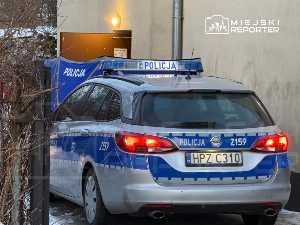 Radiowóz policyjny zaparkowany przy budynku, z niebieską taśmą zabezpieczającą w tle.