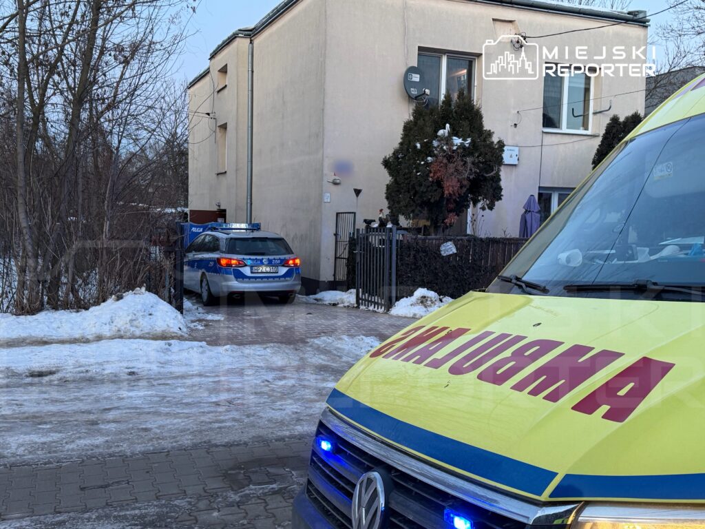 Policyjny radiowóz stoi przed budynkiem mieszkalnym, obok karetki pogotowia, na zaśnieżonym podjeździe.