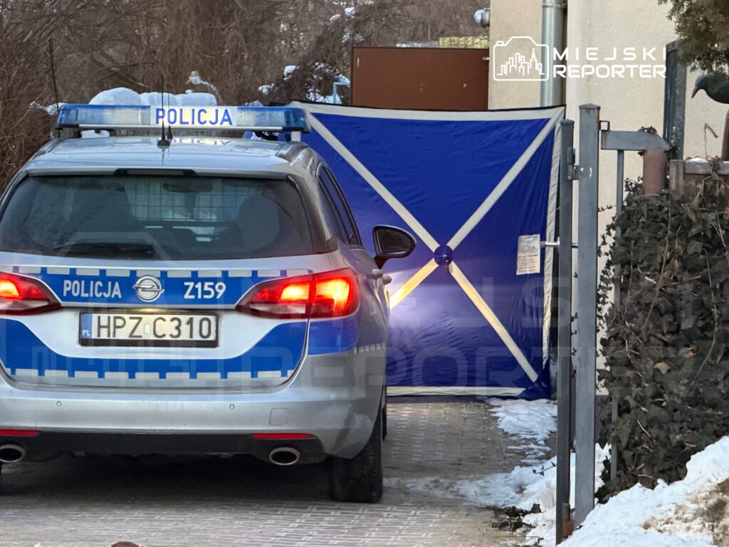 Policyjny radiowóz z włączonymi światłami stoi przed niebieską namiotową osłoną w wąskiej uliczce.