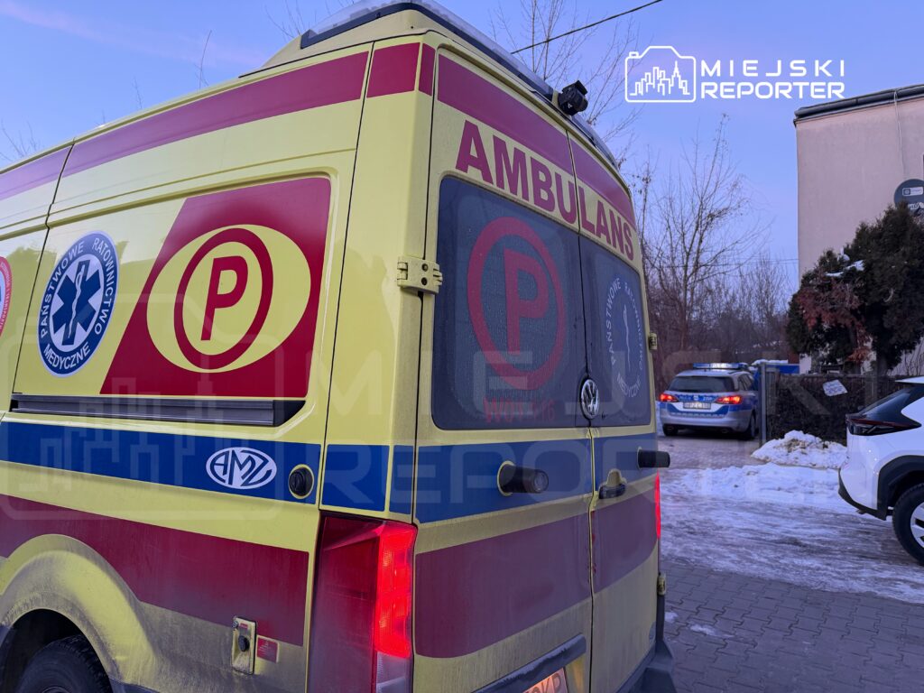 Ambulans z oznaczeniem "P" stoi na parkingu, obok widać radiowóz policyjny oraz zaparkowane samochody.