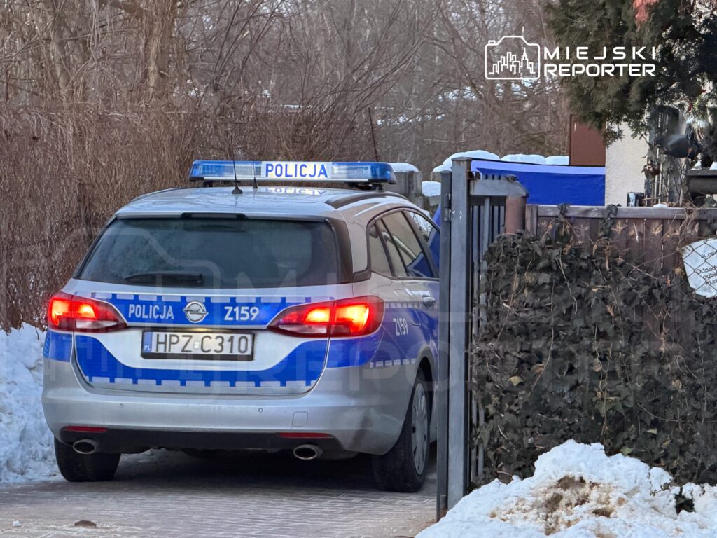 Policyjny samochód zaparkowany przy ogrodzeniu, otoczony śniegiem i zaroślami.