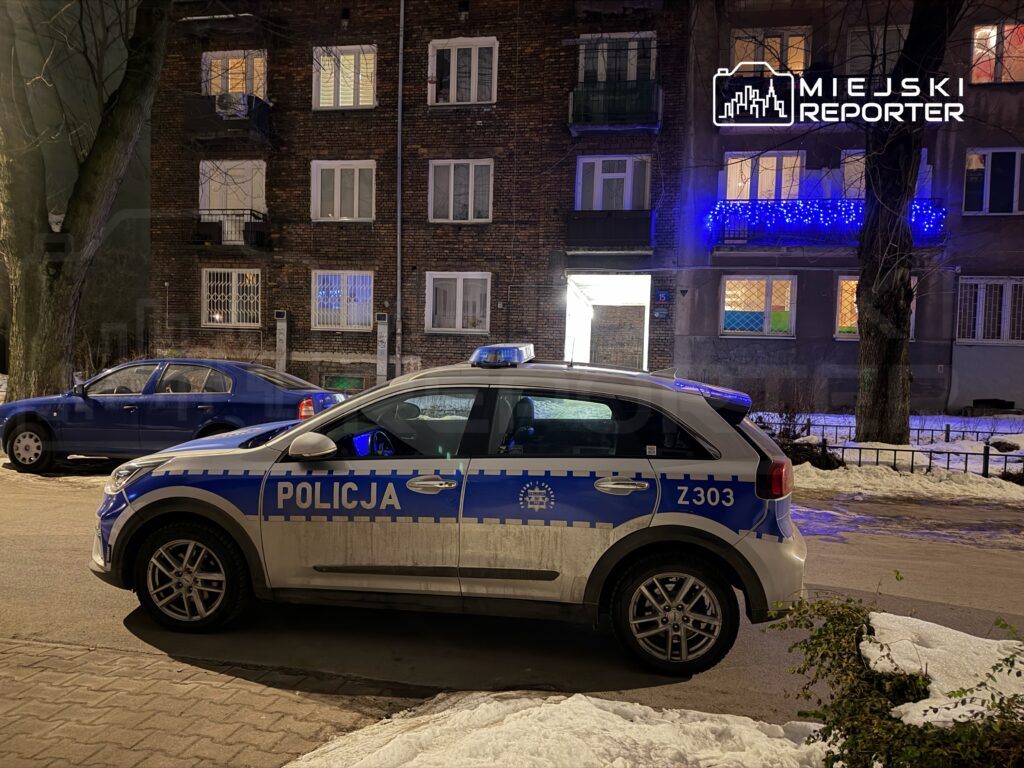 Policyjny radiowóz zaparkowany na ulicy obok budynku mieszkalnego, w tle świąteczne oświetlenie.