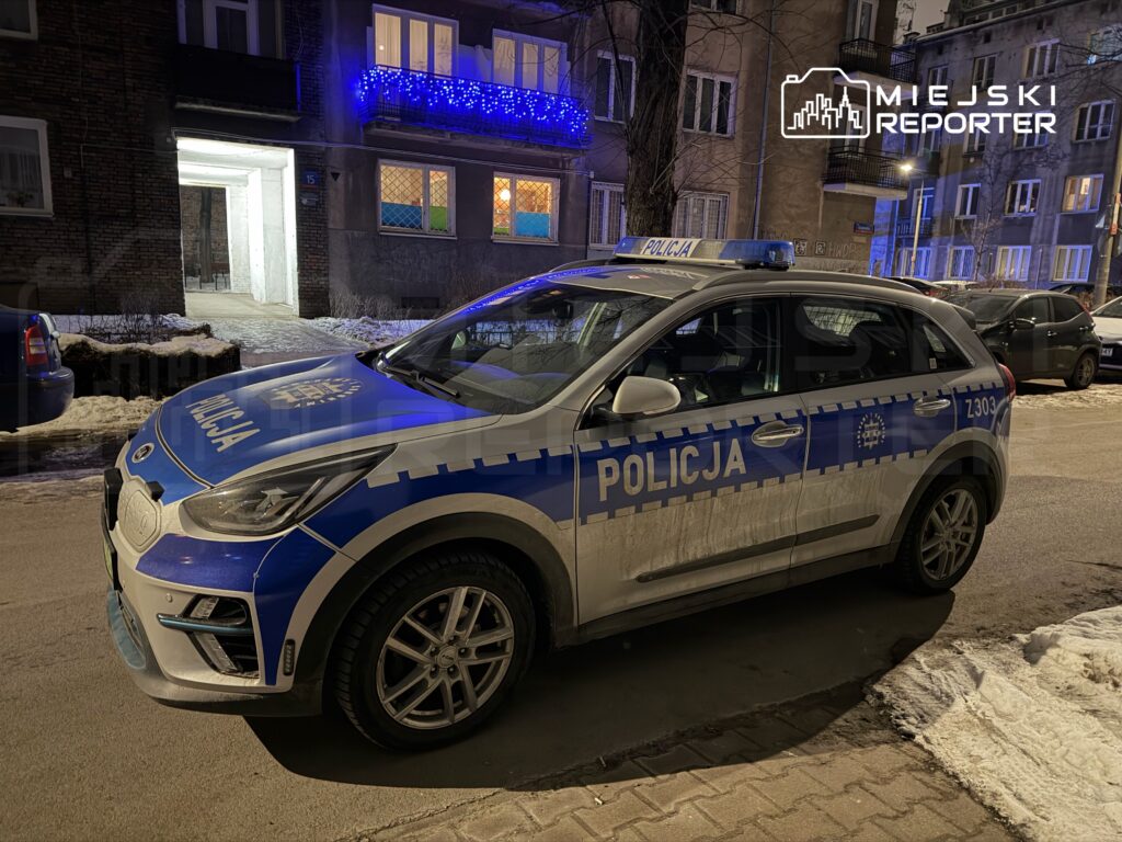 Policyjny samochód zaparkowany na oświetlonej ulicy, obok budynku z niebieskimi iluminacjami.