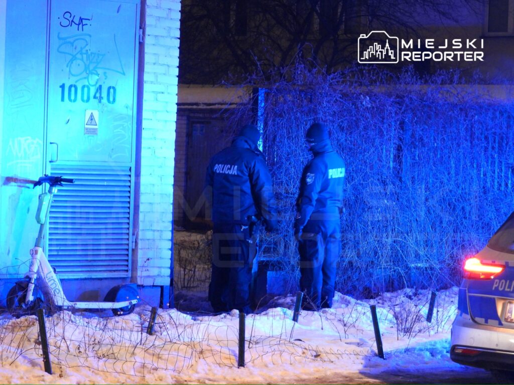 Dwóch policjantów w mundurach przeszukuje teren w niebieskim świetle radiowozu obok zaparkowanej hulajnogi.