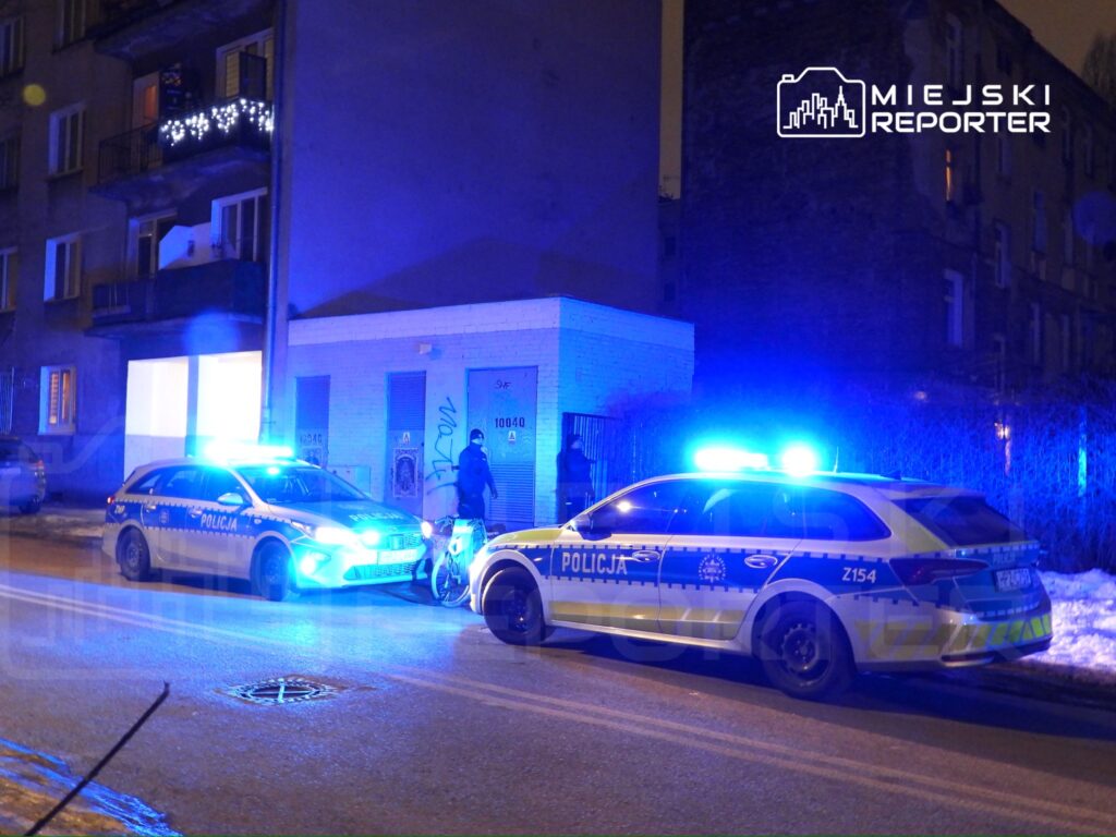 Policyjny radiowóz z niebieskimi światłami zaparkowany na ulicy, obok mężczyzny przy rowerze w nocnym otoczeniu.