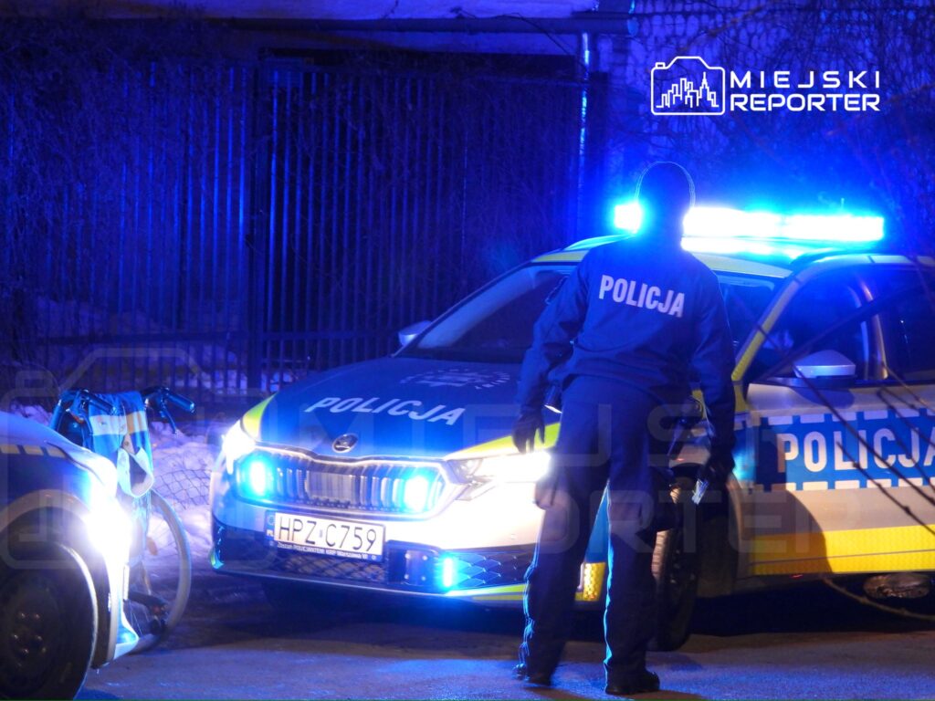 Policjant w mundurze stoi obok radiowozu z włączonymi sygnałami świetlnymi na ciemnej ulicy.