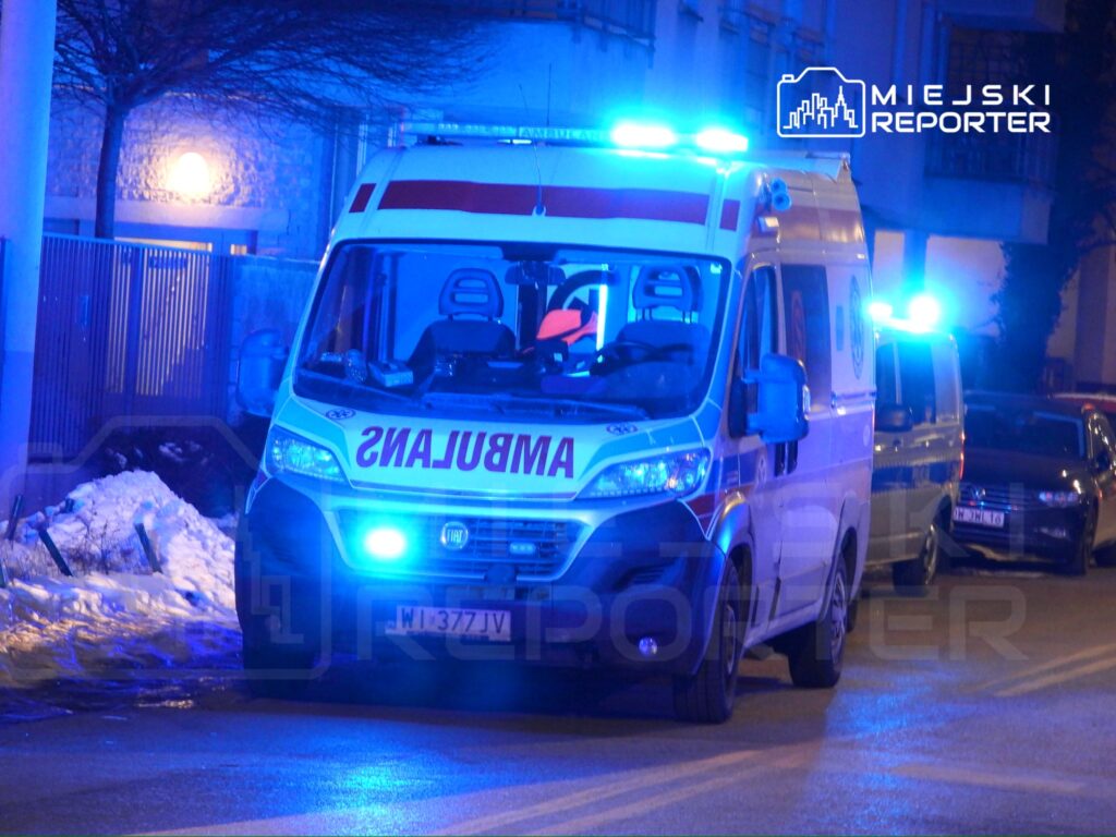 Karetka pogotowia z niebieskimi światłami zatrzymana na oświetlonej ulicy w zimowej scenerii.