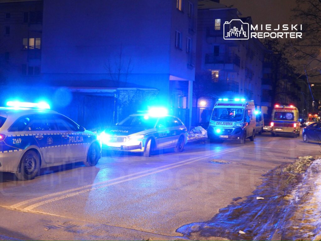 Trzy pojazdy służb ratunkowych z włączonymi sygnałami świetlnymi na nocnej ulicy, obok bloków mieszkalnych.