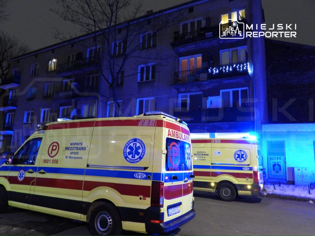 Karetki pogotowia zaparkowane przed budynkiem mieszkalnym w nocy, w tle niebieskie światła.