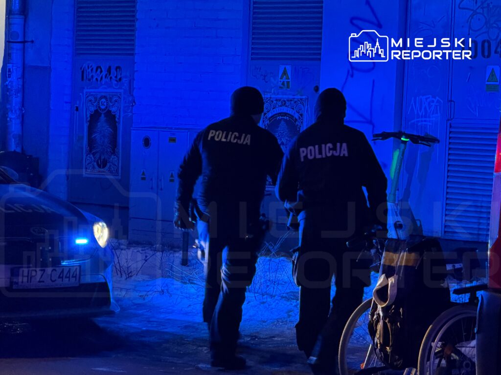 Dwaj policjanci w odblaskowych kurtkach idą w kierunku nieoświetlonej ulicy, obok stoi rower.