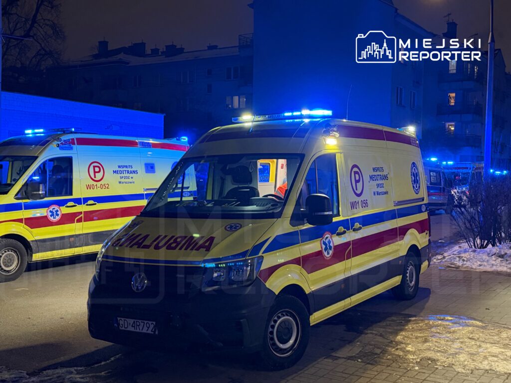 Ambulans z niebieskimi światłami zaparkowany w nocy na ulicy, w tle widać drugi ambulans oraz blok mieszkalny.