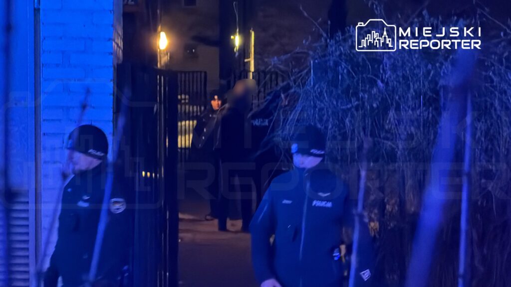 Grupa policjantów w mundurach stoi przed ogrodzeniem w oświetlonym niebieskim świetle, obserwując okolicę.