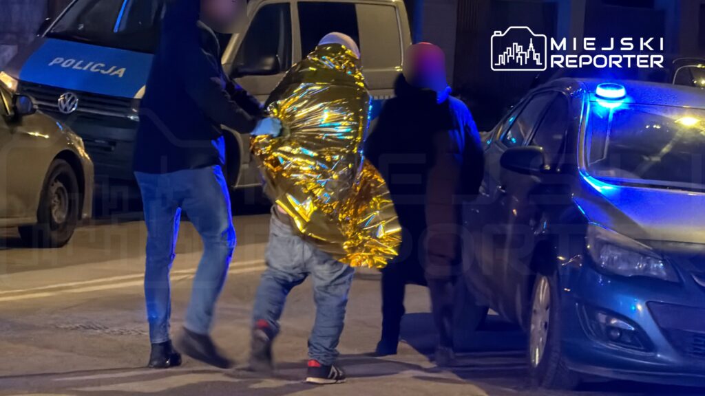Mężczyzna w złotym kocu ratunkowym prowadzony przez funkcjonariuszy policji w nocy na ulicy.