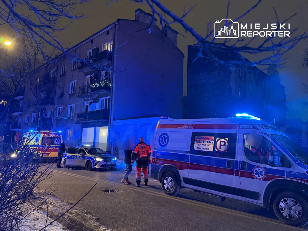 Ratownicy medyczni i policja interweniują przy bloku mieszkalnym w nocy, obok ambulansu oraz radiowozu.