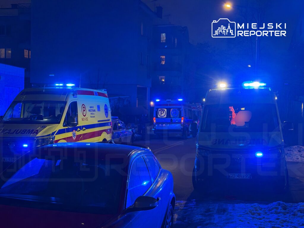 Ambulans z włączonymi sygnałami świetlnymi stoi na ulicy, obok innych pojazdów, w nocnym otoczeniu.