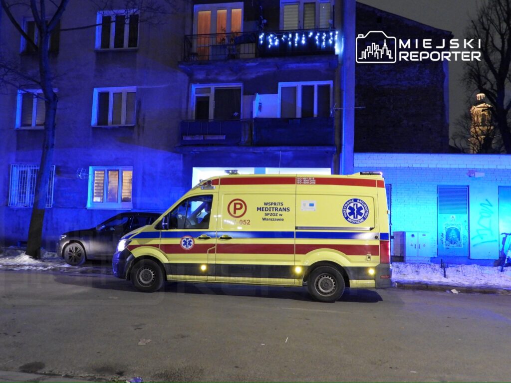 Karetka pogotowia z włączonymi sygnałami świetlnymi stoi przed budynkiem mieszkalnym oświetlonym niebieskimi światłami.