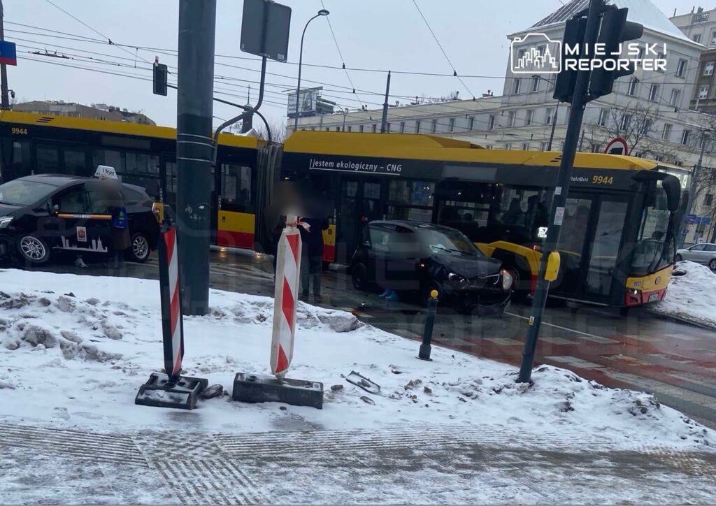 Zderzenie autobusu z samochodem osobowym na zaśnieżonej ulicy; obok stoją osoby w rozmowie.