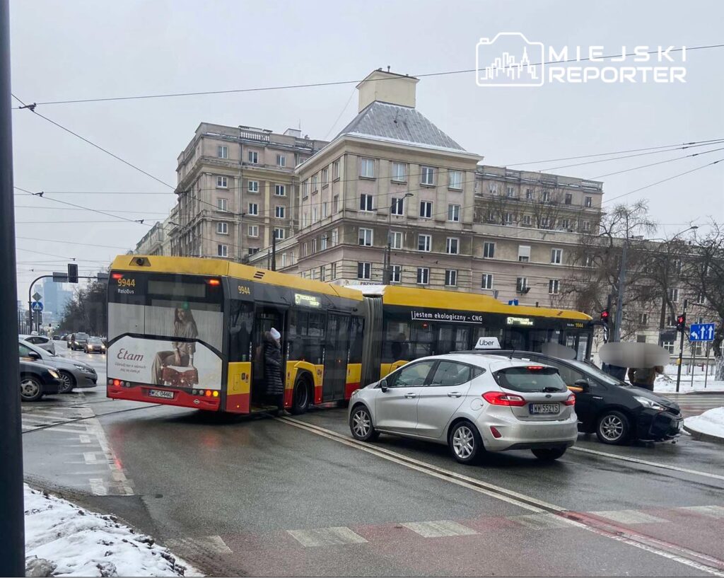 Autobus komunikacji miejskiej skręca w lewo na skrzyżowaniu, blokując ruch innych pojazdów w deszczowy dzień.