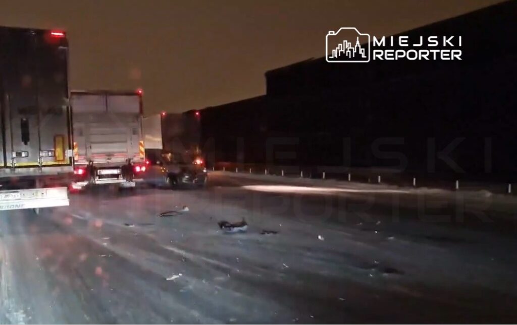 Zablokowana droga z kilkoma ciężarówkami i osobowym samochodem na zaśnieżonym odcinku, widoczna nocna sceneria.