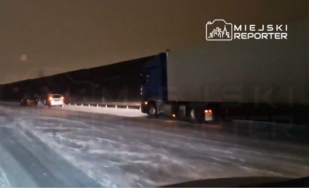 Ciężarówka porusza się po zaśnieżonej drodze w nocy, obok zatrzymanego samochodu osobowego.