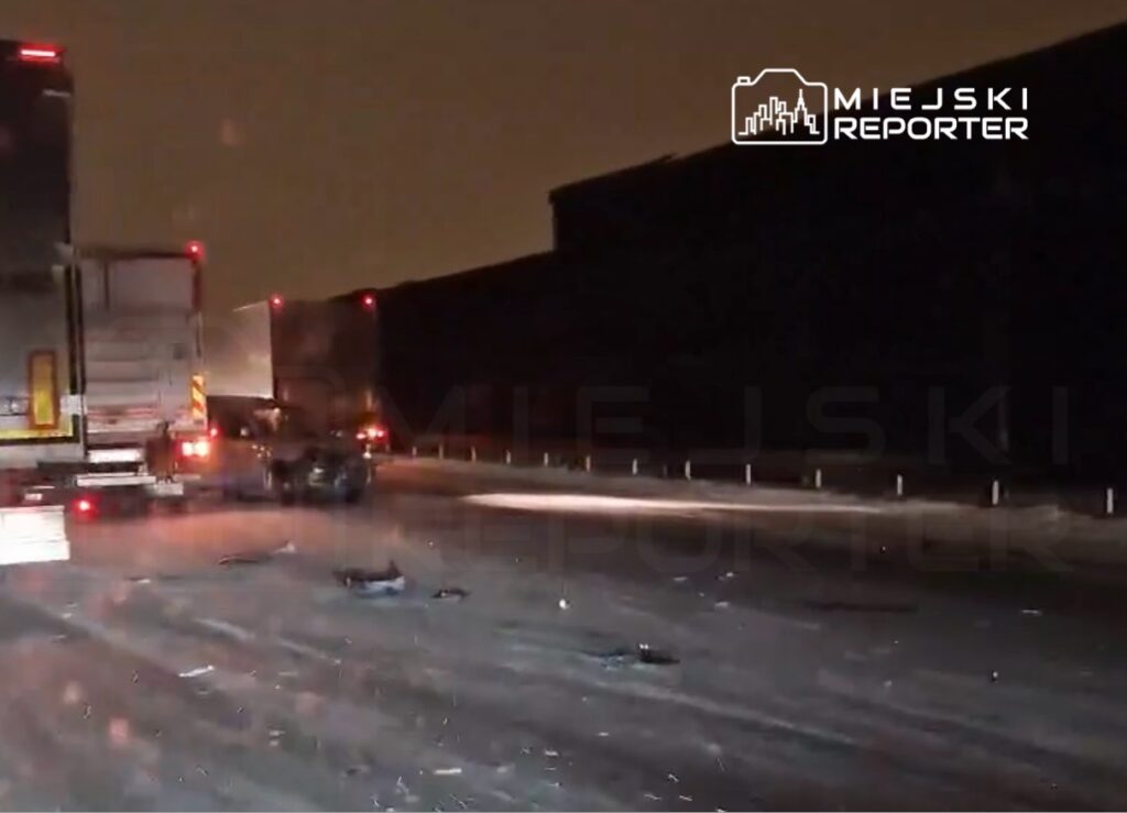 Na zaśnieżonej drodze leżą resztki zderzenia, a dwa ciężarówki stoją w ruchu, w tle ciemne niebo.