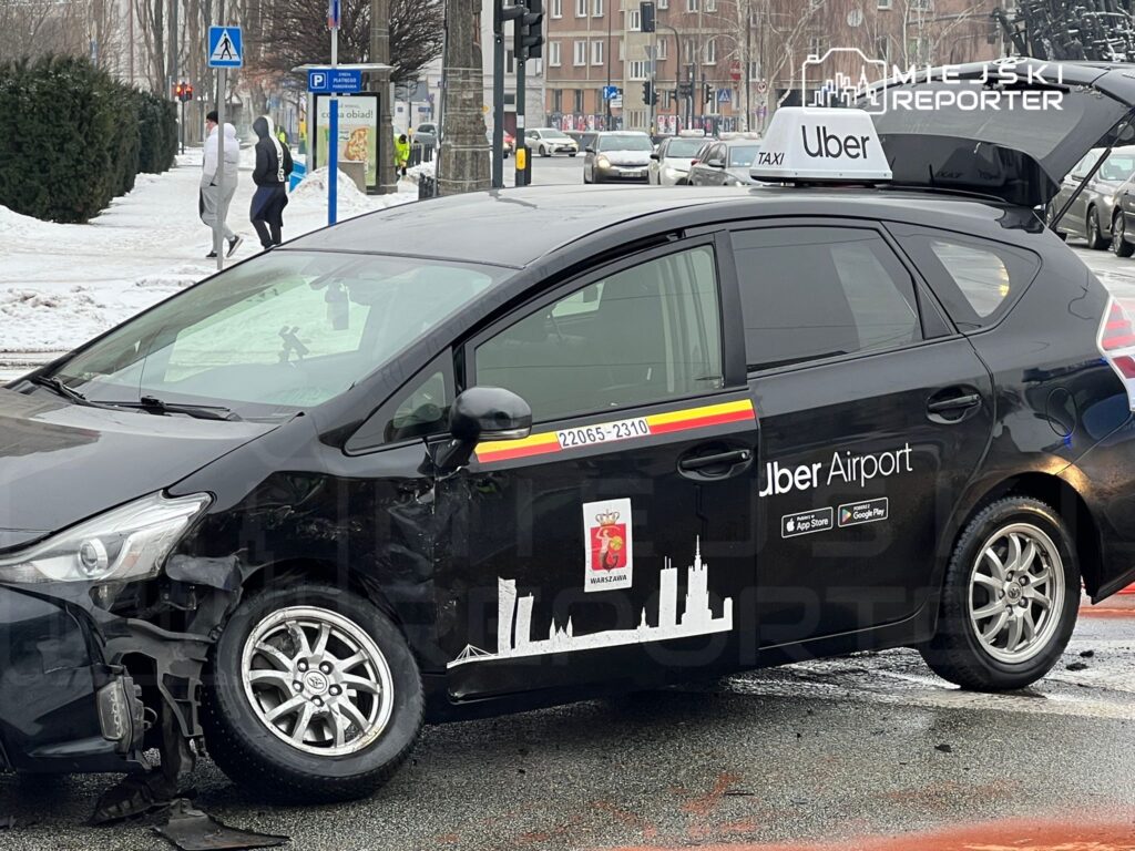 Czarny samochód osobowy z logo Uber, uszkodzony po kolizji, stoi na zaśnieżonym skrzyżowaniu w miejskim otoczeniu.