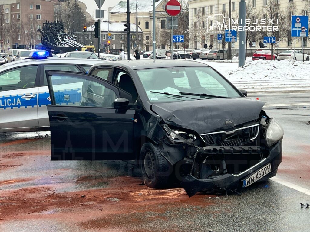 Czarny samochód osobowy z uszkodzoną przednią częścią stoi na skrzyżowaniu, obok policyjny radiowóz z włączonymi sygnałami.