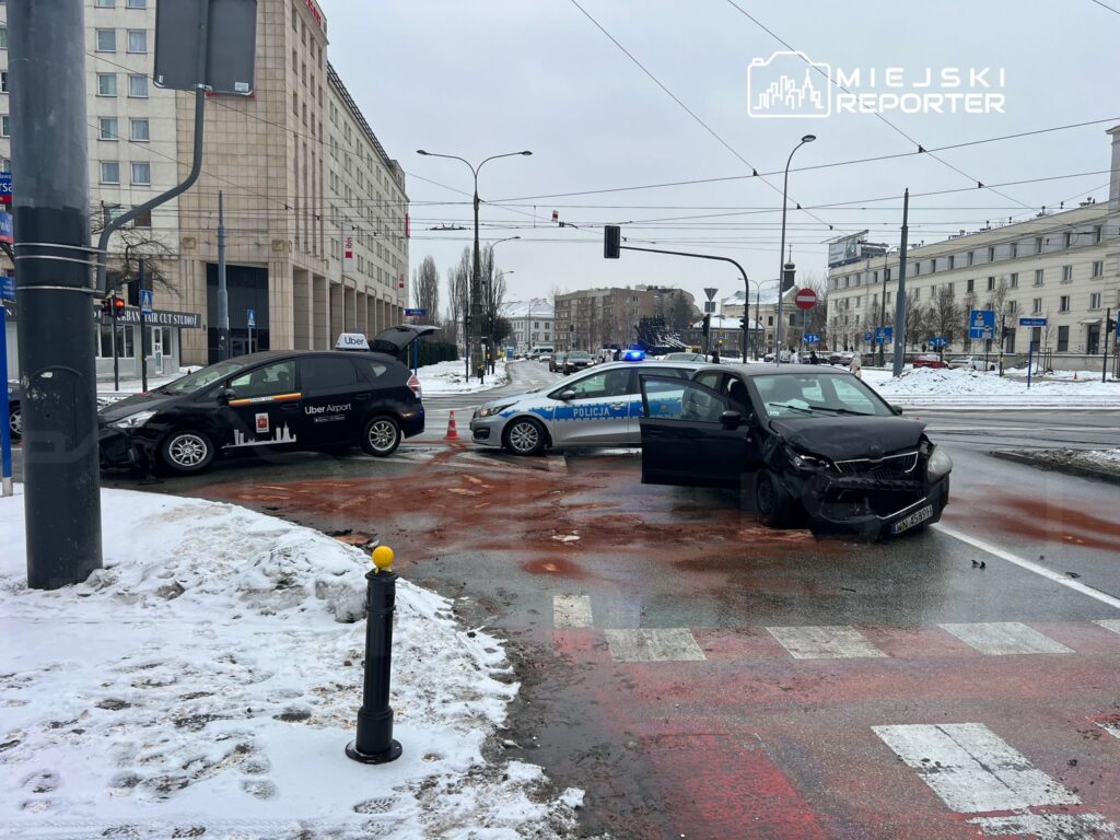 Dwa uszkodzone samochody osobowe na skrzyżowaniu, jeden z logo Ubera, a obok radiowóz policji. Na jezdni widoczne plamy płynów.