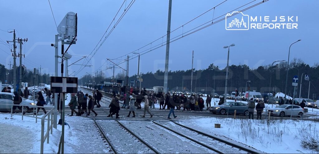 Tłum ludzi przechodzi przez tory kolejowe w zaśnieżonym otoczeniu, obok zaparkowanych samochodów.