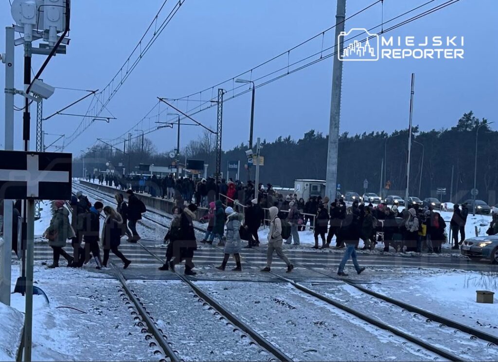Tłum ludzi przechodzi przez tory kolejowe w zimowej scenerii, w tle widoczny przystanek i samochody.