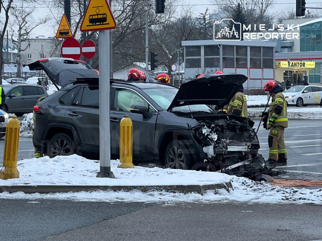 Czarny samochód osobowy z uszkodzoną przednią częścią, obok strażacy w hełmach sprawdzający sytuację na skrzyżowaniu.