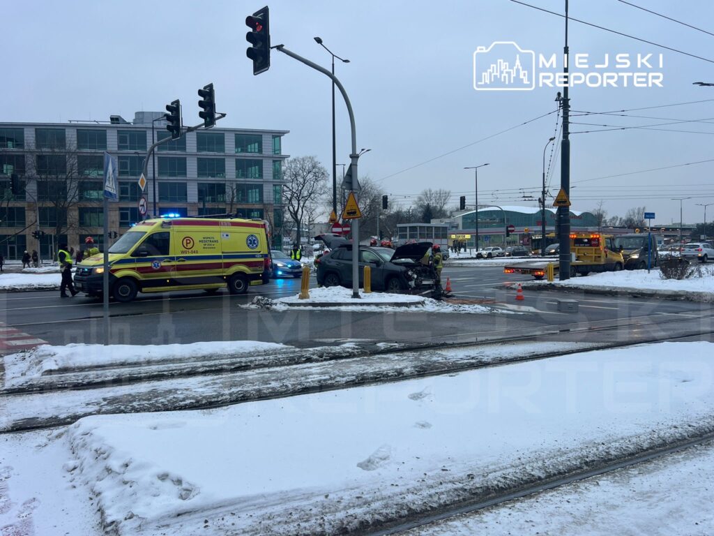 Karetka pogotowia oraz policjanci zabezpieczają miejsce wypadku drogowego na skrzyżowaniu w zimowej scenerii.