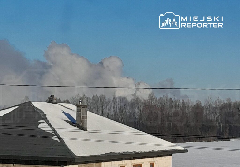 Biały dym unoszący się nad zadrzewionym terenem, w tle niebieskie niebo i pokryty śniegiem dach budynku.