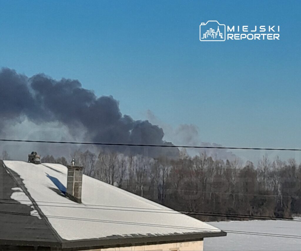 Czarny dym unoszący się nad lasem, widoczny z dachu budynku pokrytego śniegiem w słoneczny dzień.