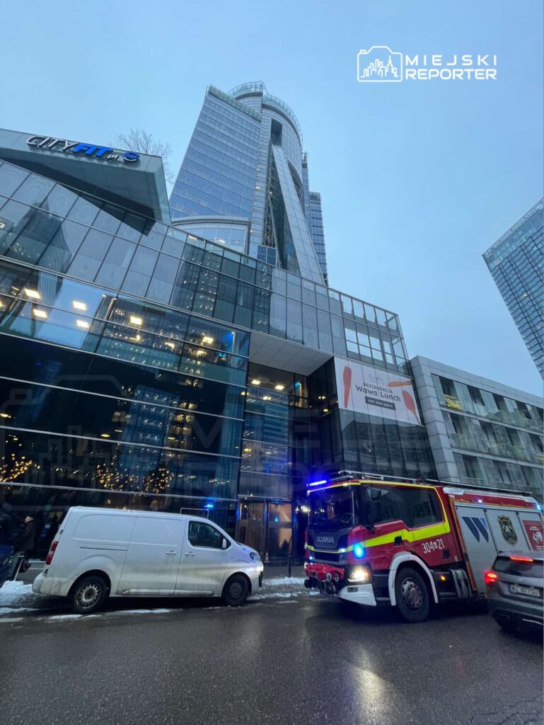 Straż pożarna z włączonymi sygnałami świetlnymi stoi przed nowoczesnym budynkiem z przeszkloną fasadą.