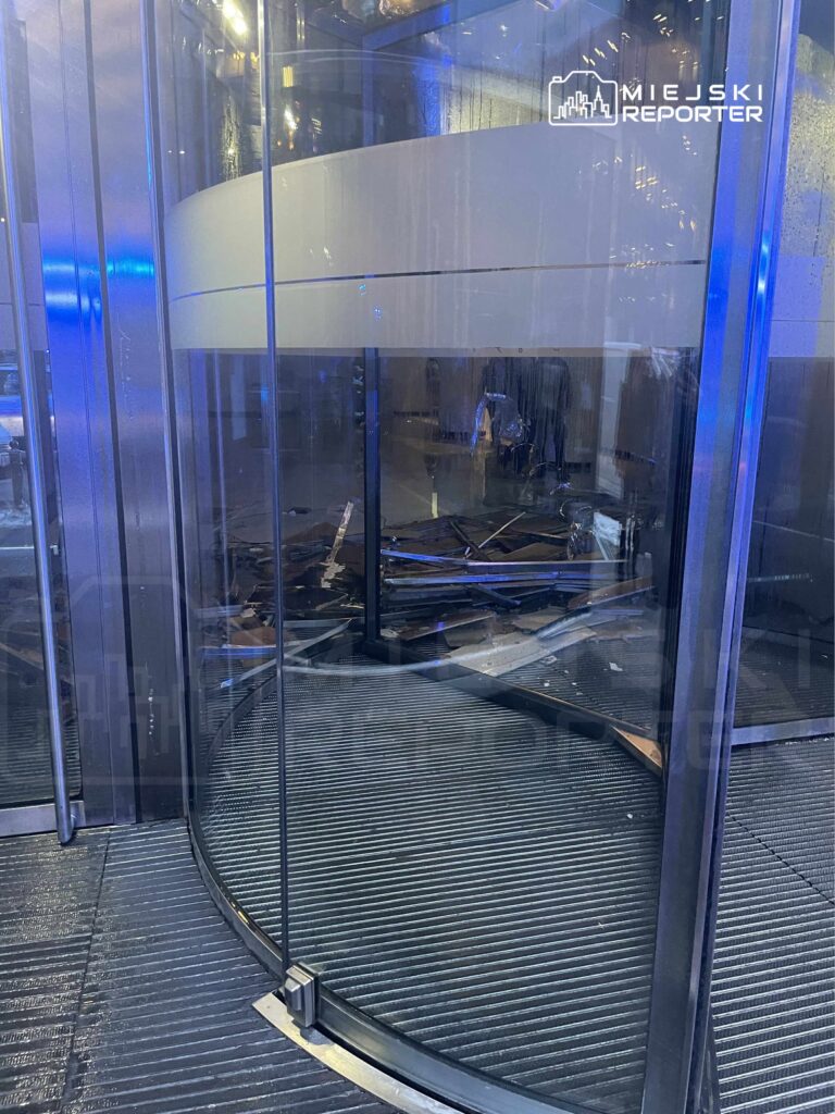Zniszczona szklana winda w nowoczesnym biurowcu z porozrzucanymi odpadami na podłodze.
