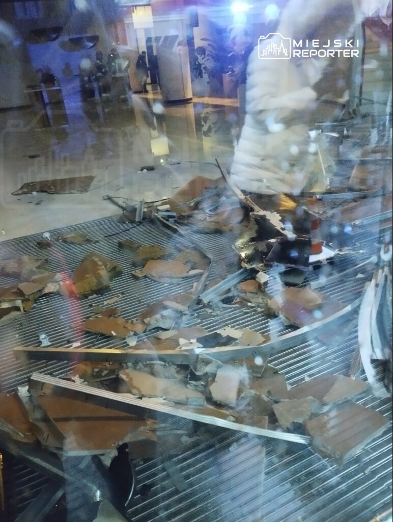 Na podłodze widać rozbite fragmenty szkła i gruz, obok stoi osoba w białej kurtce, w tle widoczni strażacy.