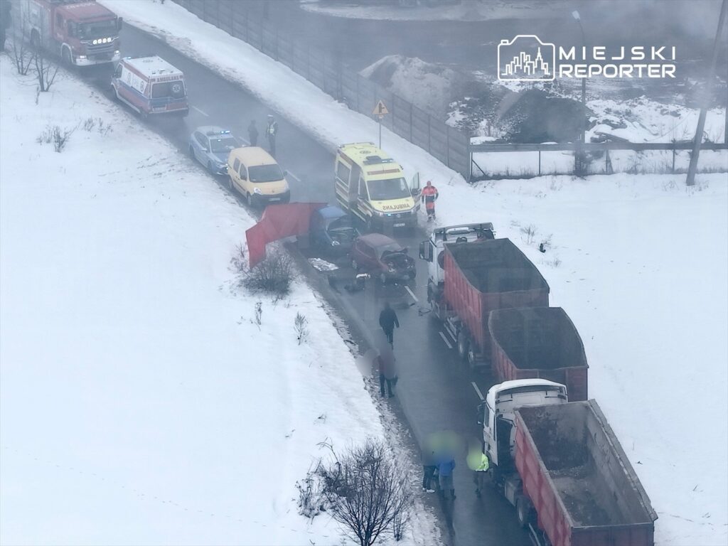 Na zaśnieżonej drodze doszło do wypadku, w którym uczestniczyły dwa pojazdy. W tle widać karetki i radiowóz, a obok grupę ludzi.