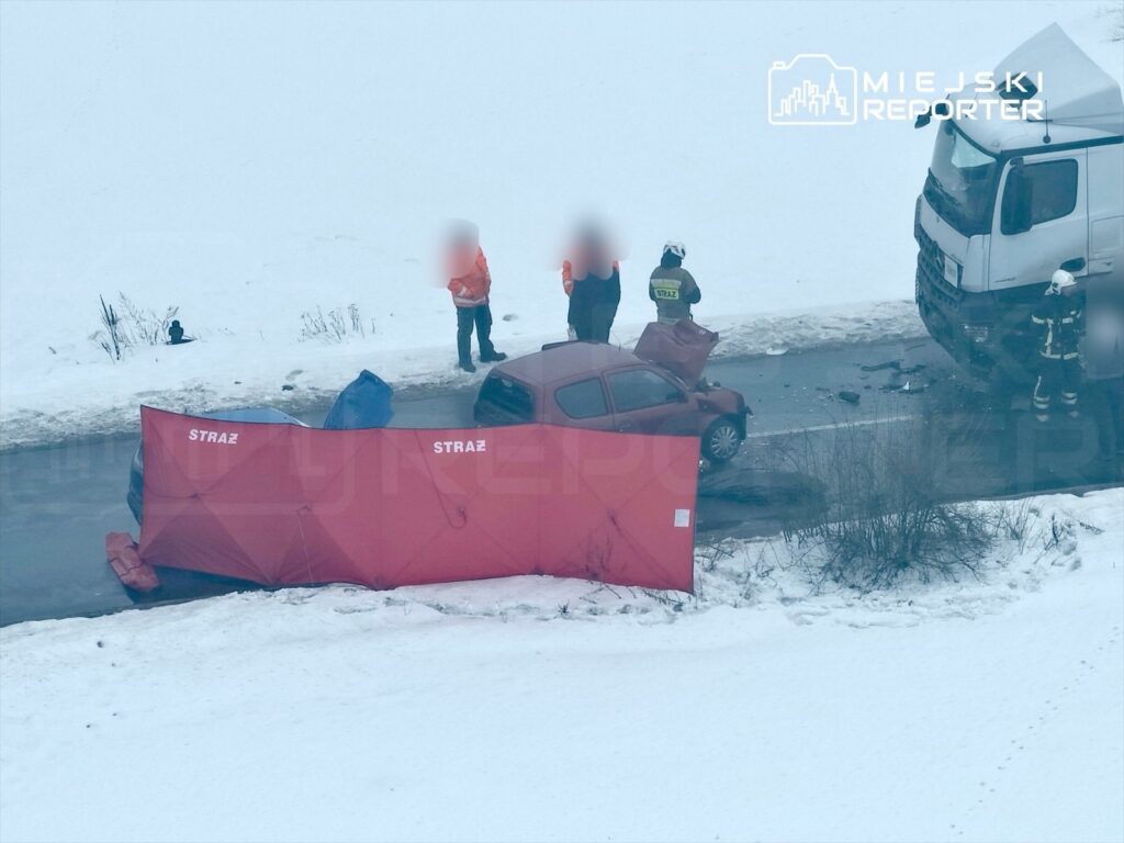 Na drodze po wypadku stoją strażacy, obok uszkodzony samochód osobowy i ciężarówka, wszystko w zimowej scenerii.